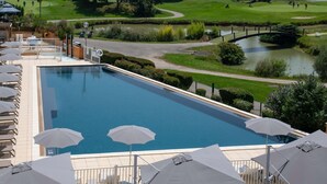 Una piscina con sillones de descanso y sombrillas, un campo de golf al fondo y un puente sobre un estanque.
