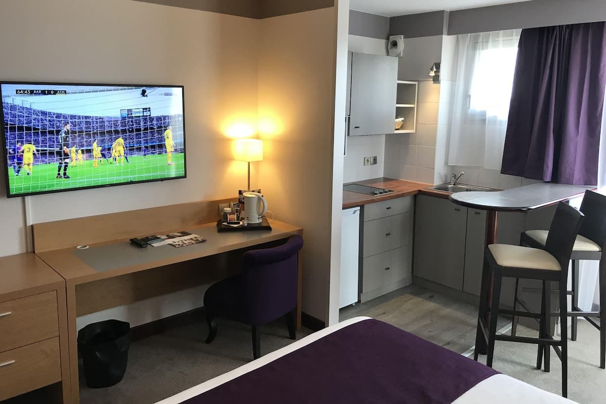 A hotel room with a TV, desk, chair, bed, and kitchenette.