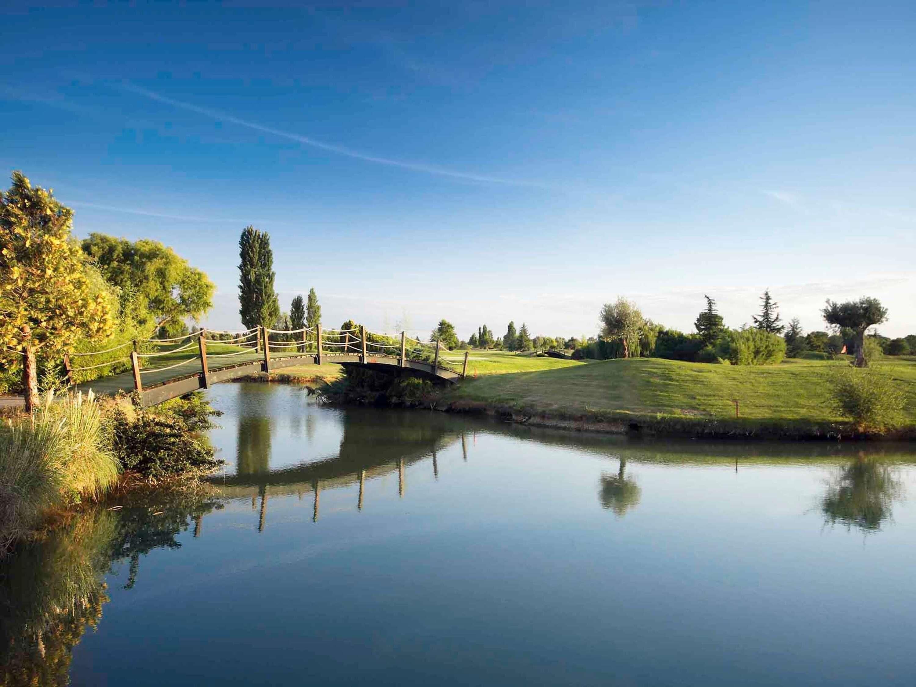 Una scena tranquilla lungo il fiume, con un ponte di legno, acqua calma e vegetazione rigogliosa.