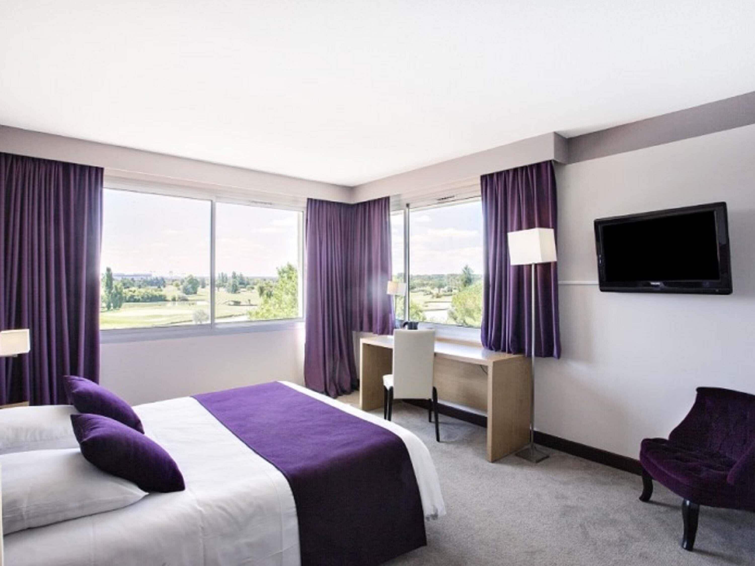 A hotel room with a bed, desk, chair, TV, and large windows with curtains.