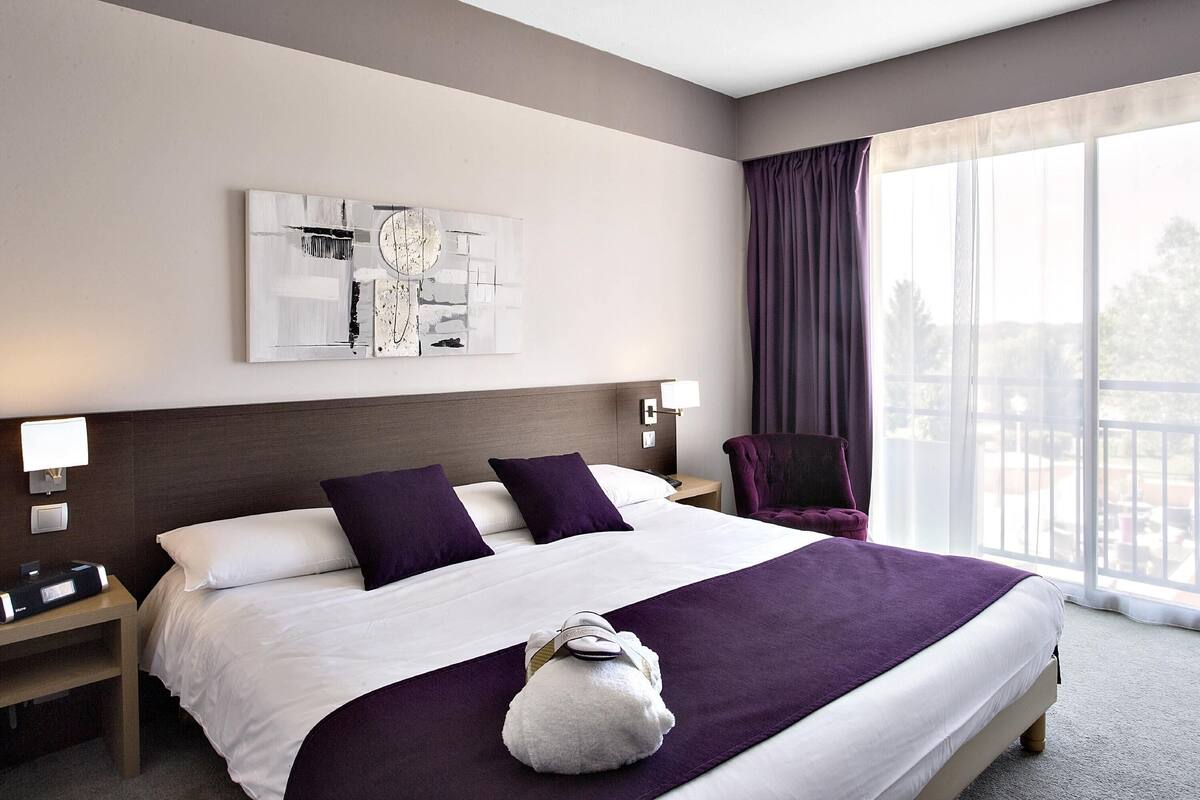 Une chambre d’hôtel avec un grand lit, une tête de lit, une table de chevet avec une lampe, une chaise et une fenêtre avec des rideaux.