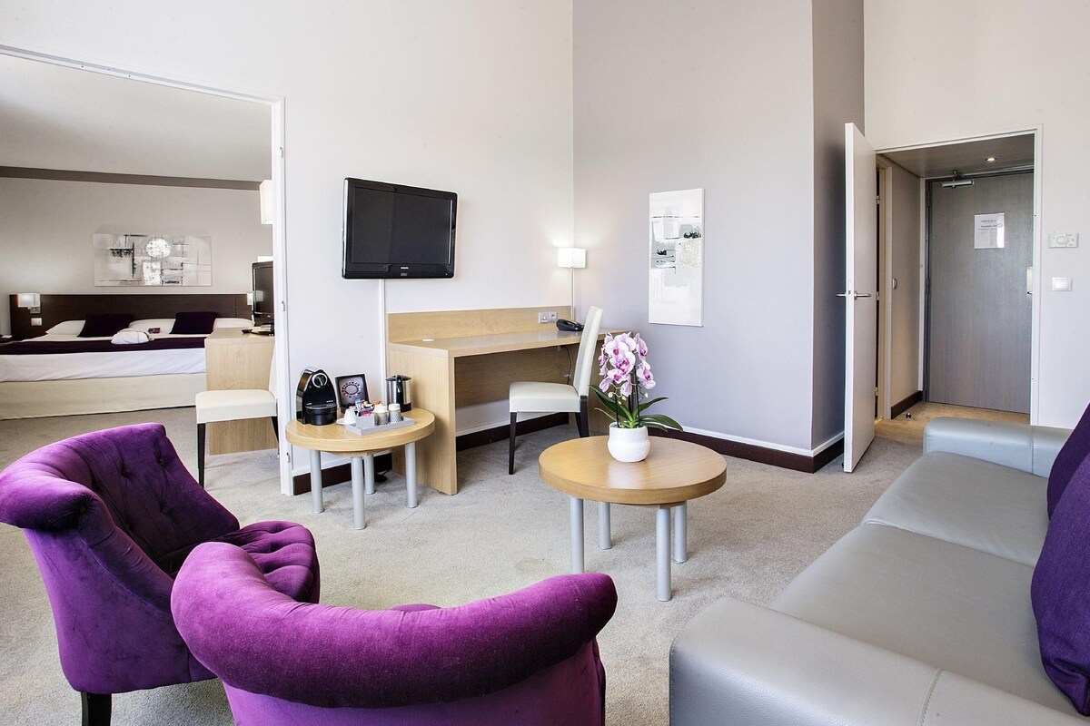 A hotel room with a bed, a desk, a television, a small table with a vase of flowers, and purple armchairs.