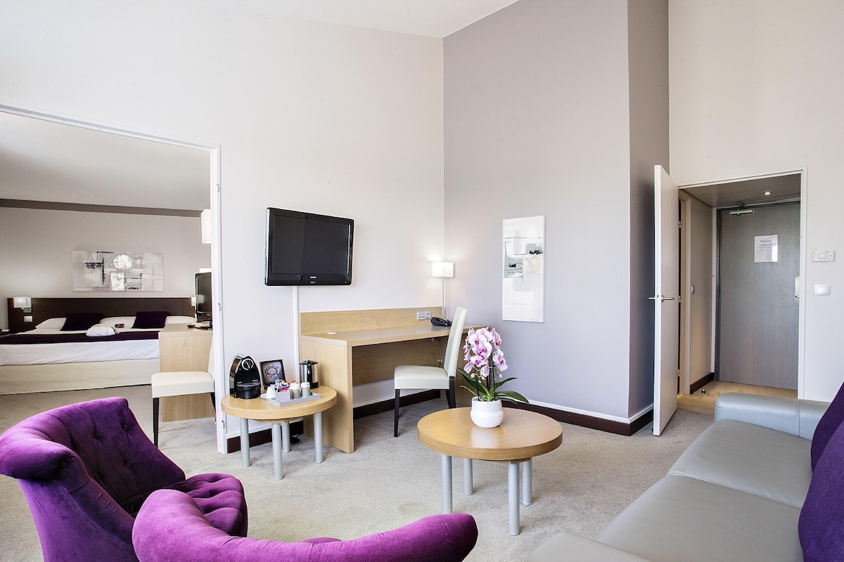 A hotel room with a bed, a desk, a television, a small table with a vase of flowers, and two purple armchairs.