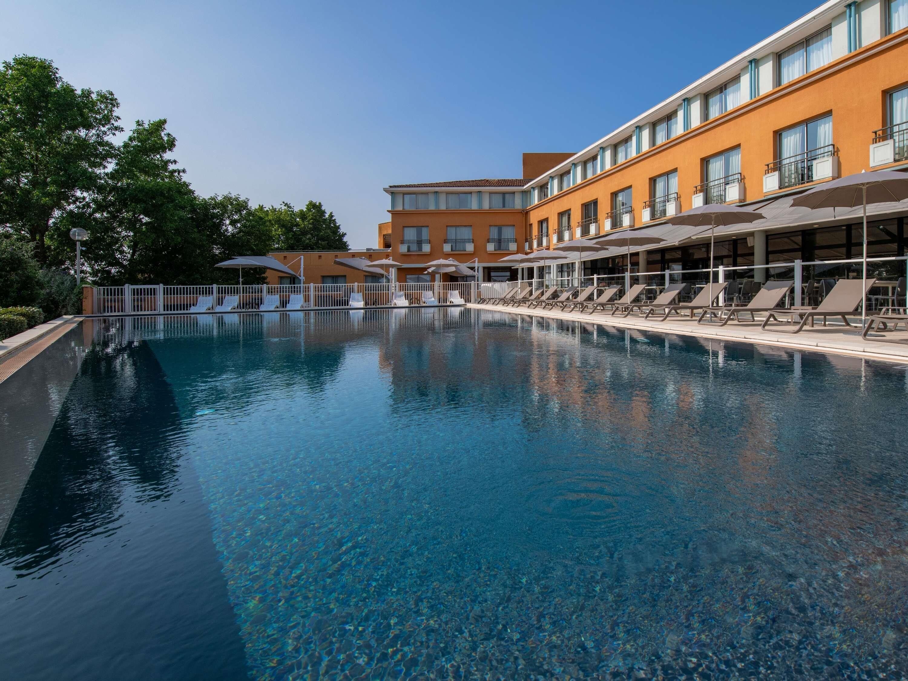 A swimming pool with lounge chairs and umbrellas, surrounded by a building with multiple floors and balconies.