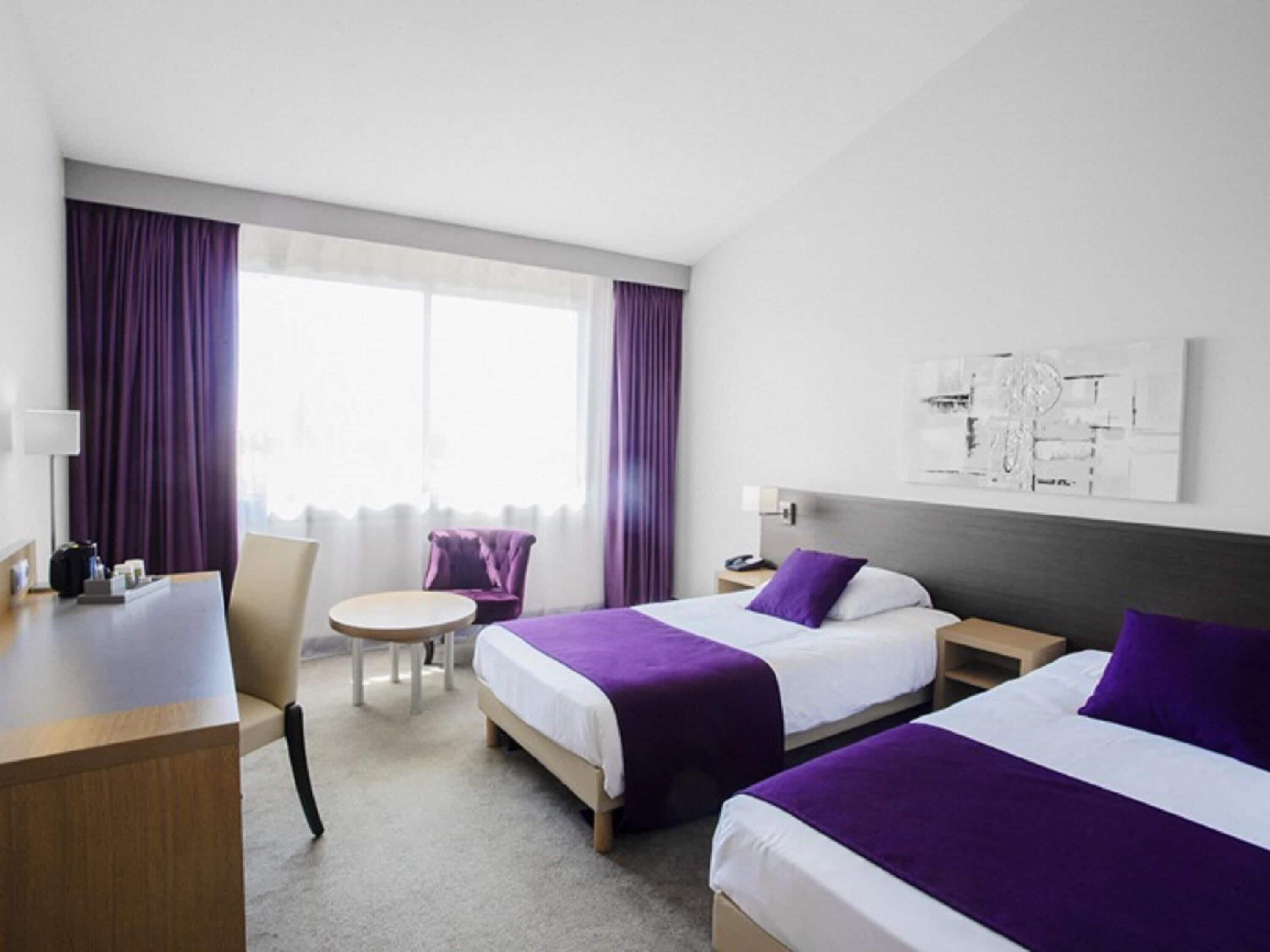 A hotel room with two beds, a desk, a chair, a small table, and a window with curtains.
