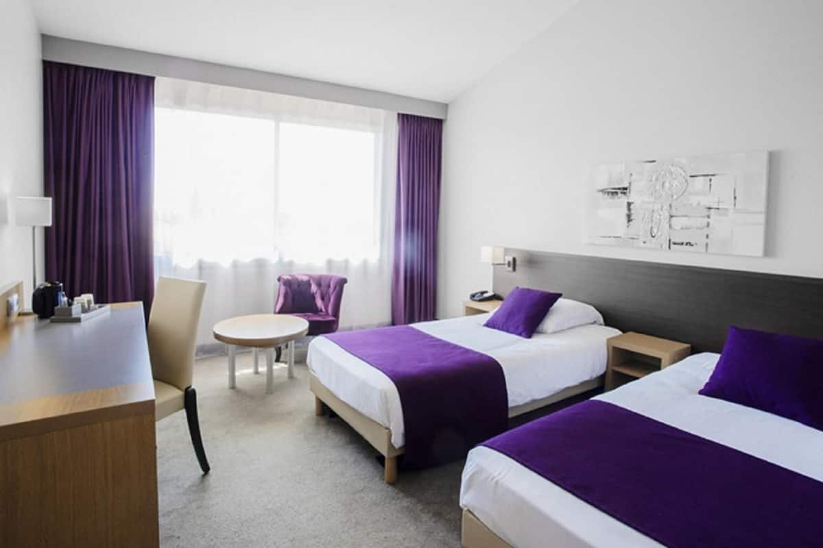 A hotel room with two beds, a desk, a chair, a small table, and a window with curtains.