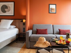 A modern hotel room with a grey sofa, a white bed, and a small table with fruit and drinks. - Mercure Toulouse Aeroport Golf de Seilh (Seilh)