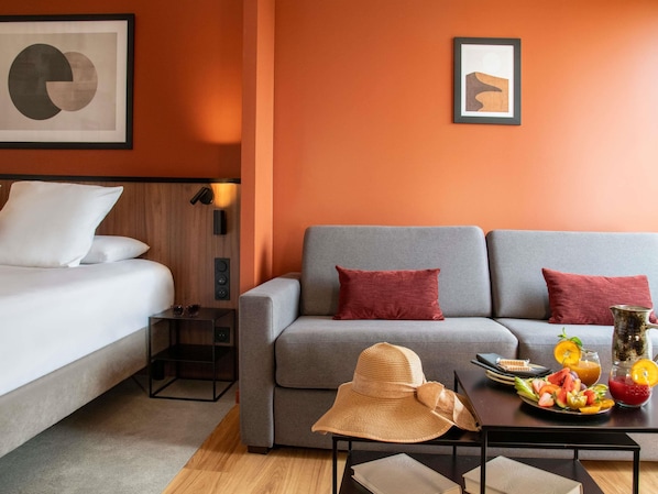 A modern hotel room with a grey sofa, a white bed, and a small table with fruit and drinks.