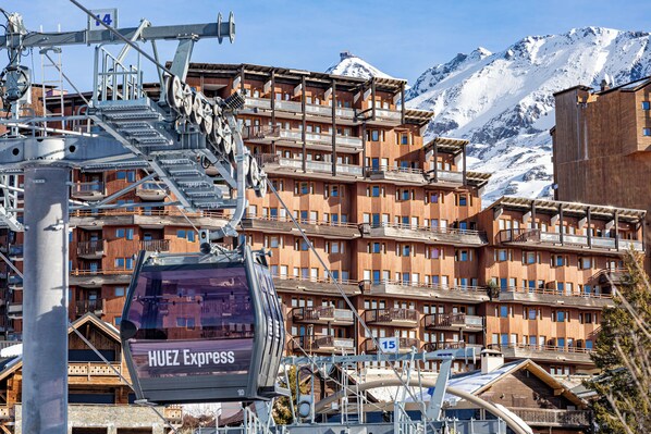 Un hotel de varios pisos con balcones, un telesilla y montañas nevadas al fondo.