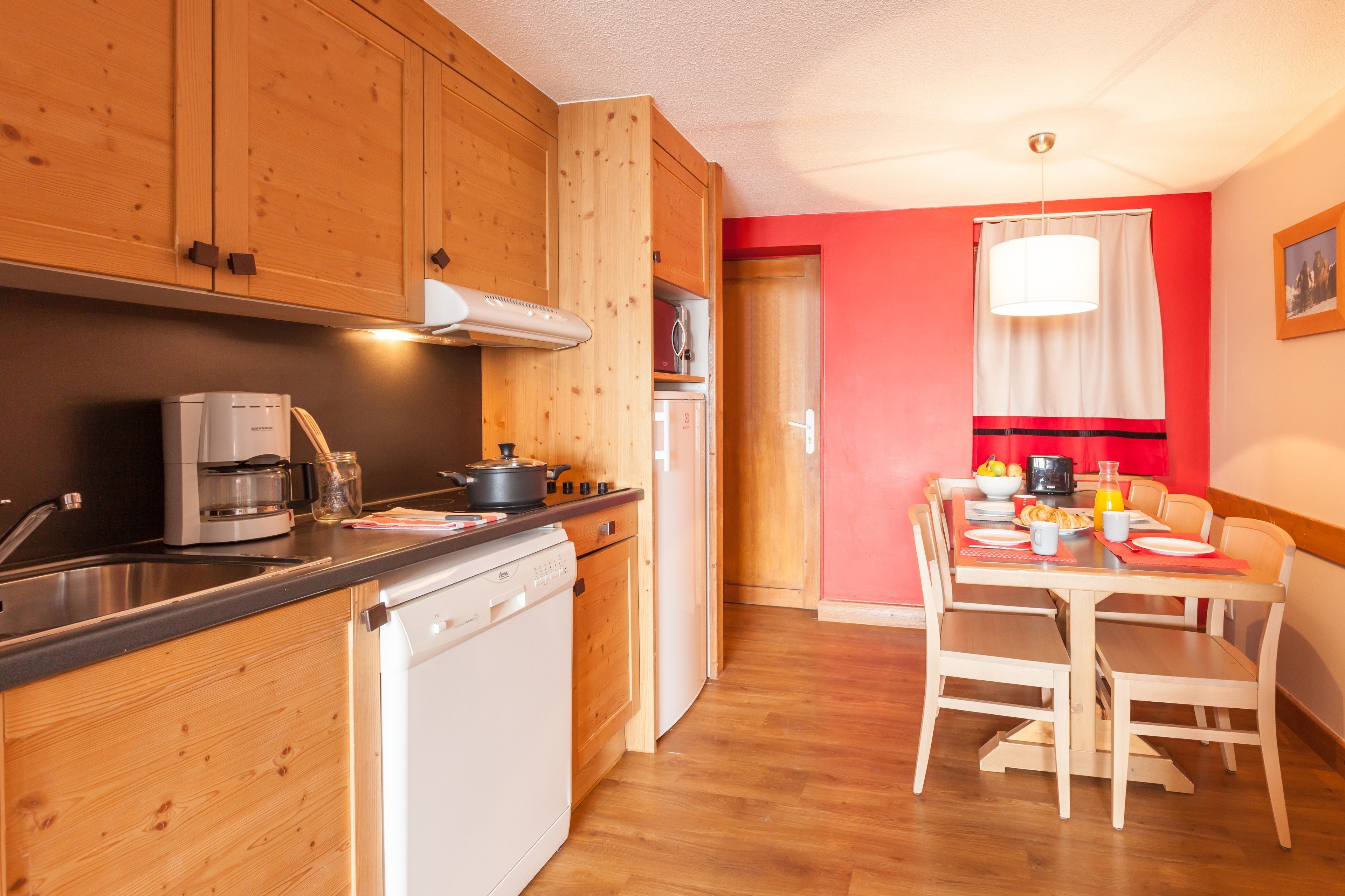 A compact kitchen with wooden cabinets, a dishwasher, a coffee maker, and a dining area with a table and chairs.