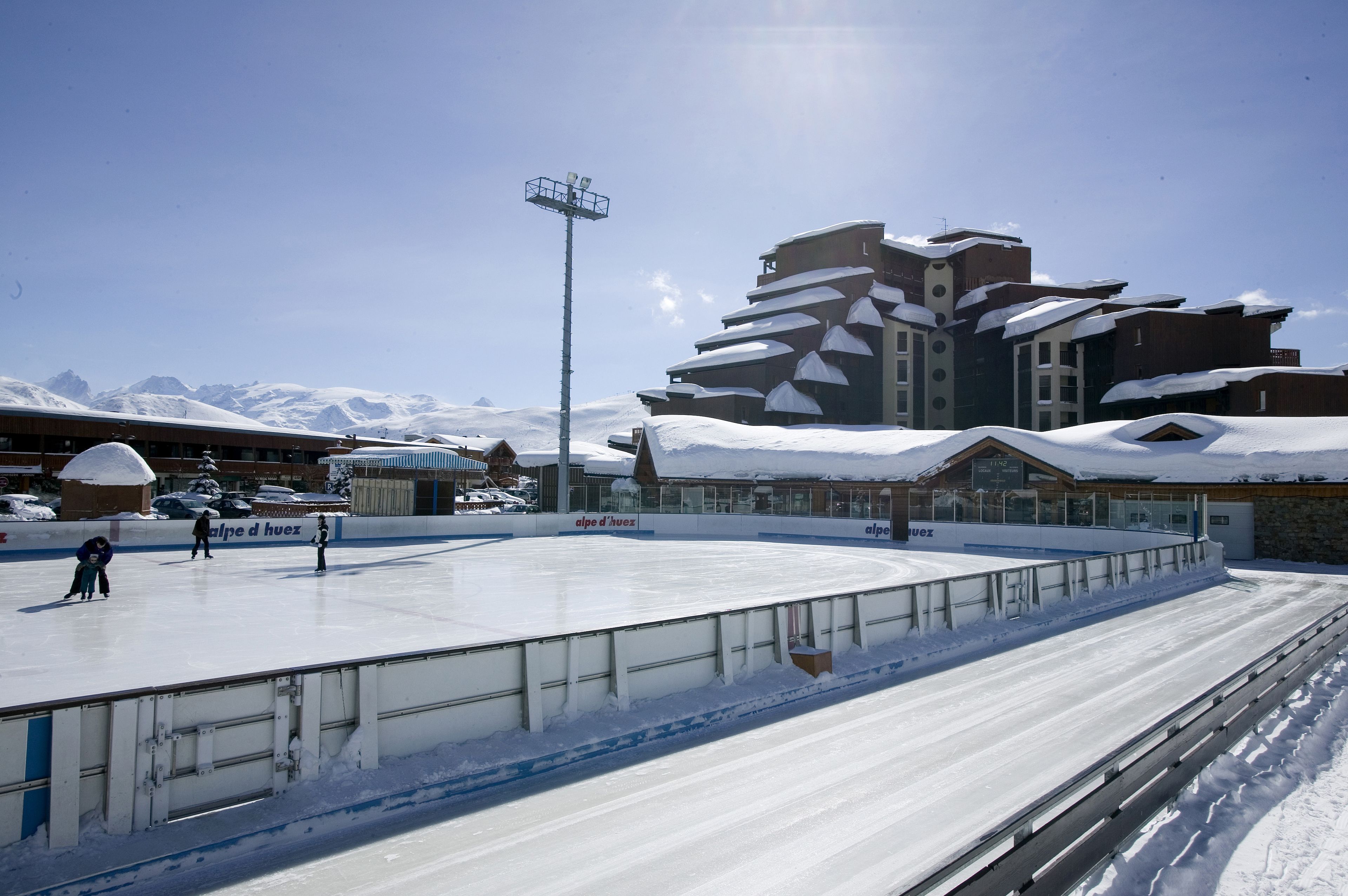An outdoor ice skating rink with a clear sky, surrounded by buildings with snow-covered roofs.