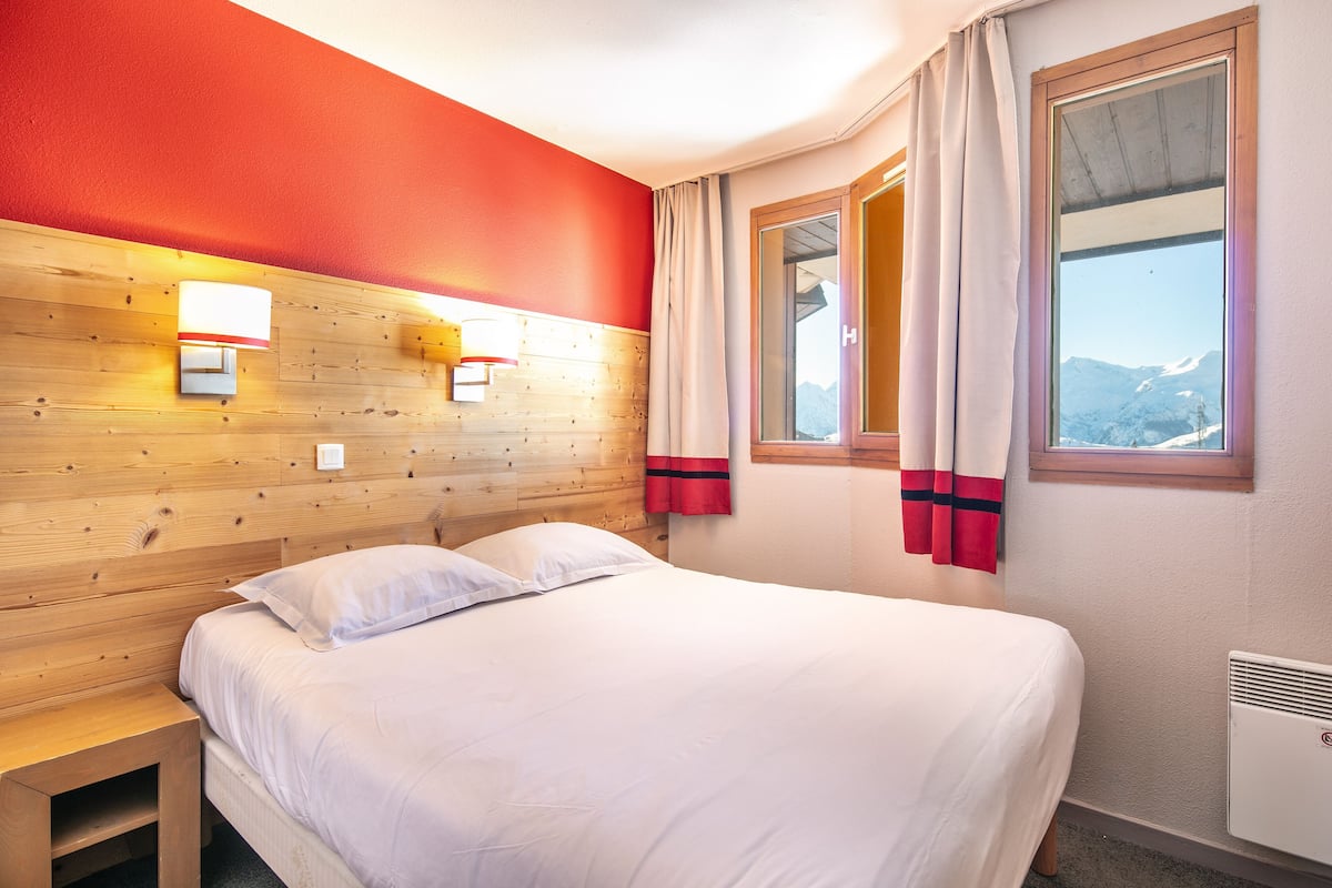 A hotel room with a wooden headboard, a bed with white linens, a nightstand, a window with curtains, and a view of mountains.