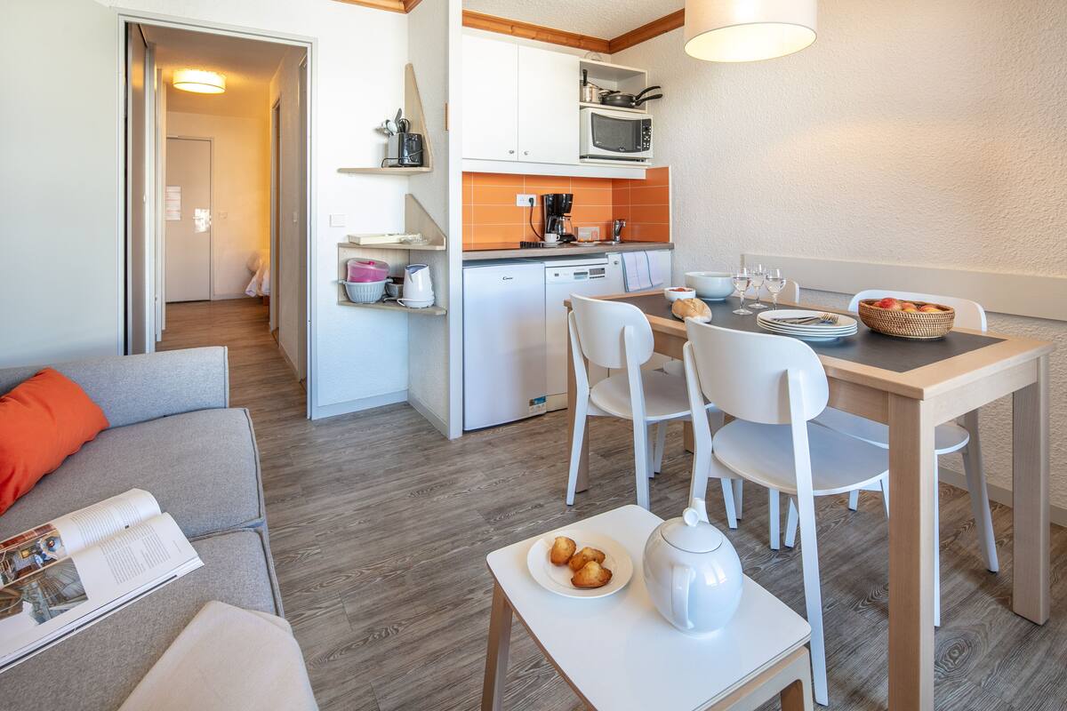 A modern kitchen with white cabinets, a microwave, and a coffee maker. There is a dining table with chairs and a small white table with a teapot and a plate of pastries.