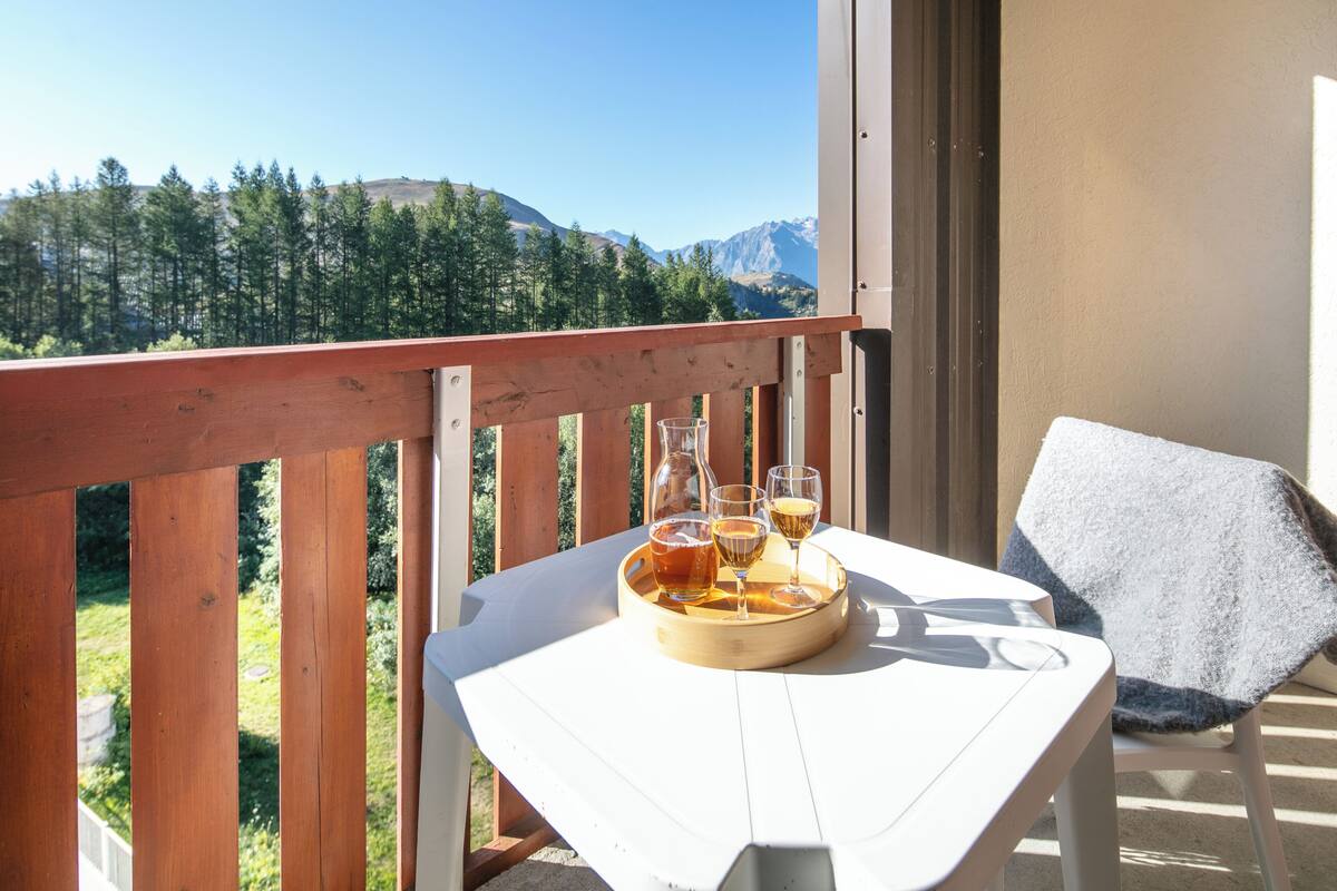 A balcony with a table holding a tray with glasses and a bottle, a chair with a towel, and a mountain view.