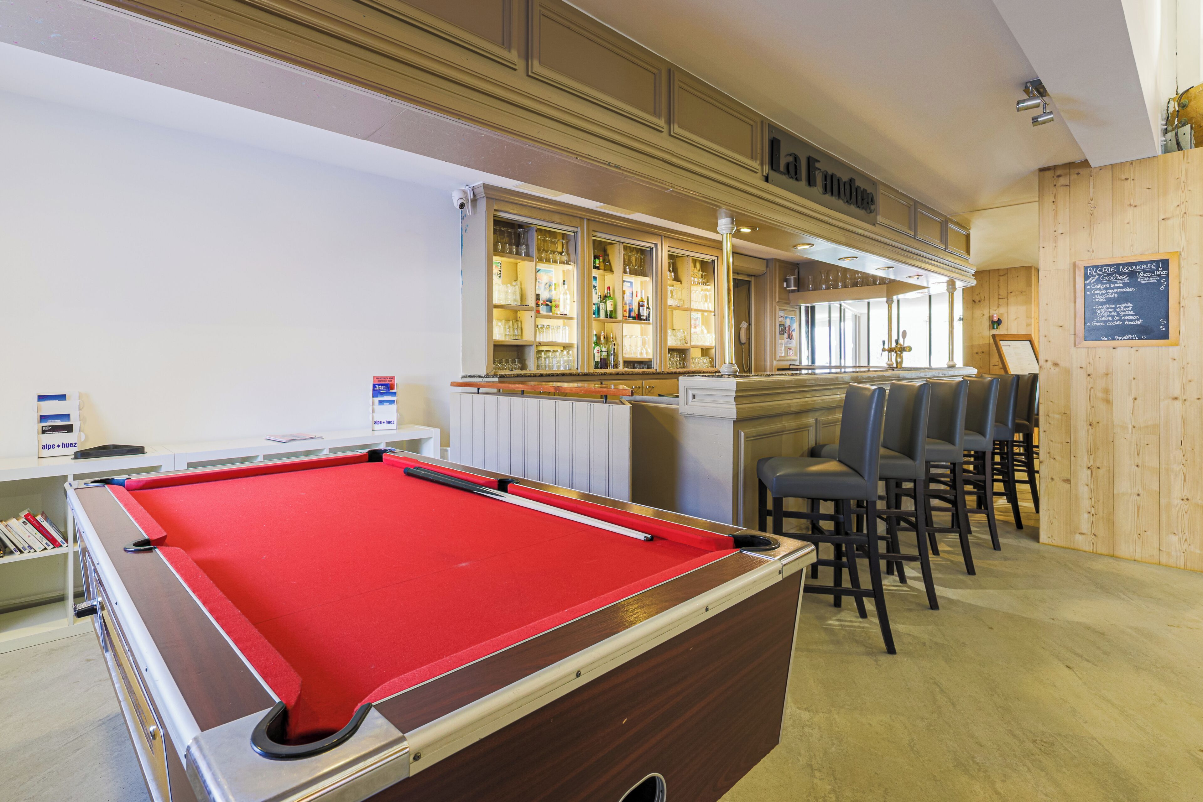 Une table de billard recouverte d’un drap de feutre rouge, un coin bar avec un comptoir en bois et des tabourets, et un menu écrit à la craie sur le mur.