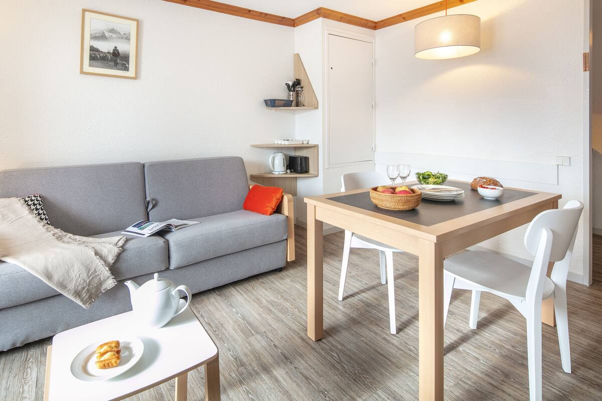 A modern living room with a grey sofa, a wooden dining table with chairs, a coffee table with a teapot and croissant, and a bookshelf with decorative items.