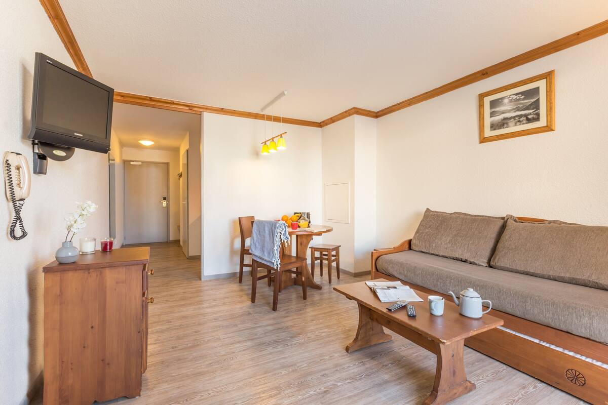 A living room with a wooden sofa, a wooden coffee table with a teapot and cups, a TV mounted on the wall, and a dining area with a table and chairs.