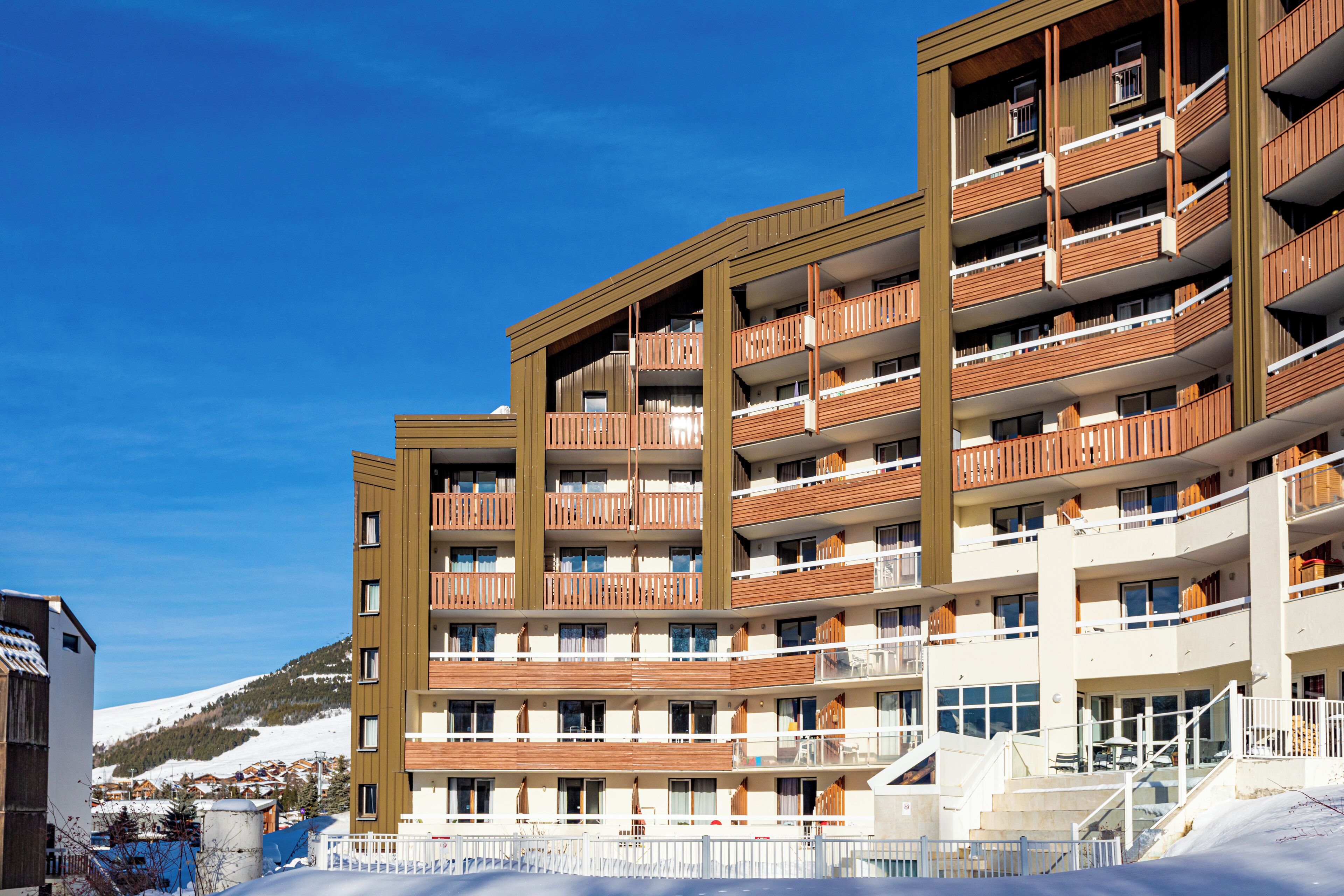 A multi-story hotel with balconies, a ski slope in the background, and a clear blue sky.