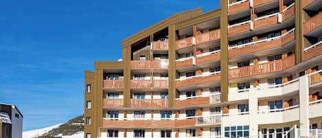 A multi-story hotel with balconies, a ski slope in the background, and a clear blue sky.