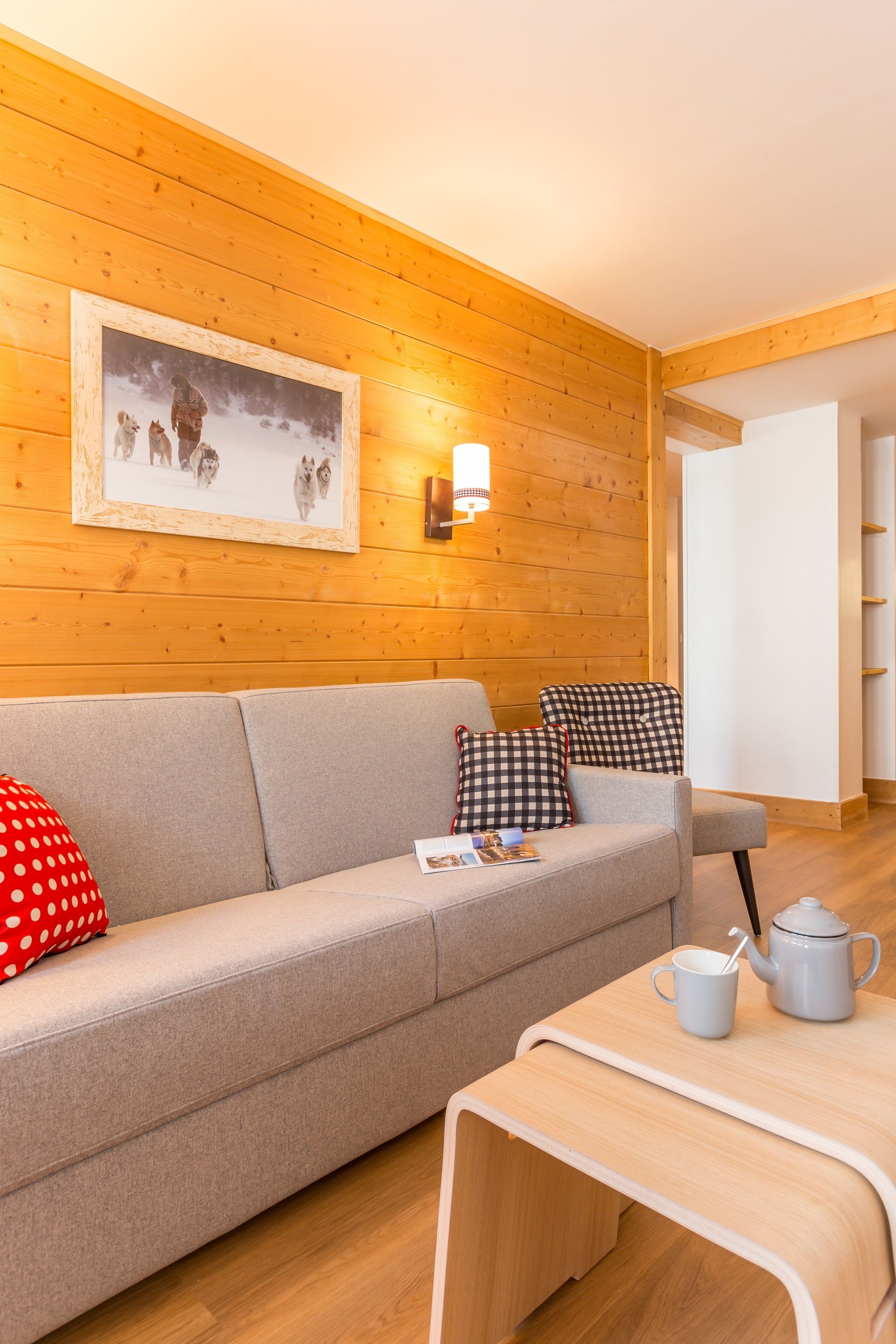 Una habitación acogedora con pared de madera, un sofá gris con cojines cuadriculados, una mesita con tetera y taza, y un cuadro en la pared.