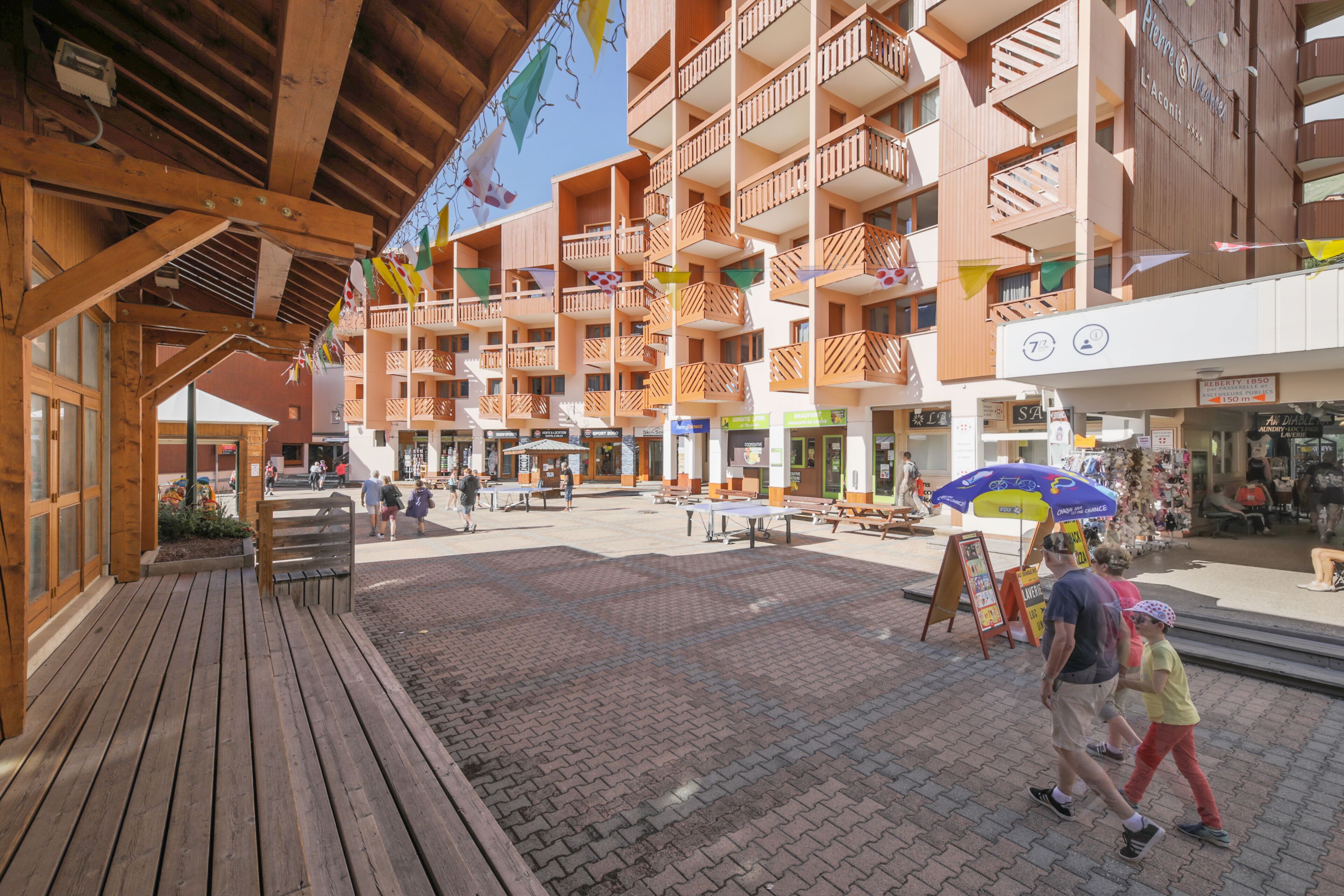 Una terraza de madera con un patio de ladrillo, un área de estar cubierta y un edificio de varios pisos con balcones y tiendas.