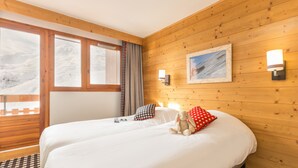 A hotel room with a wooden interior, a large bed with white bedding, a view of a snowy landscape through the window, and a framed picture on the wall. - Pierre & Vacances Residence Aconit (Les Belleville)