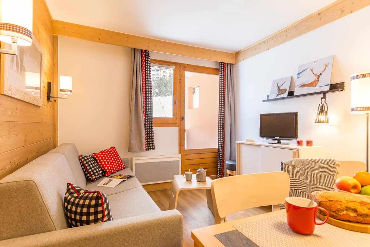 Una habitación acogedora con sofá, una mesa de comedor con hogaza de pan y una taza roja, televisor y ventana con cortinas.