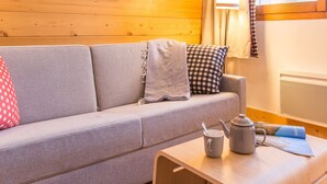 A cozy room with a wooden wall, a grey sofa, a small table with a kettle and a book, and a framed picture on the wall. - Pierre & Vacances Residence Aconit (Les Belleville)