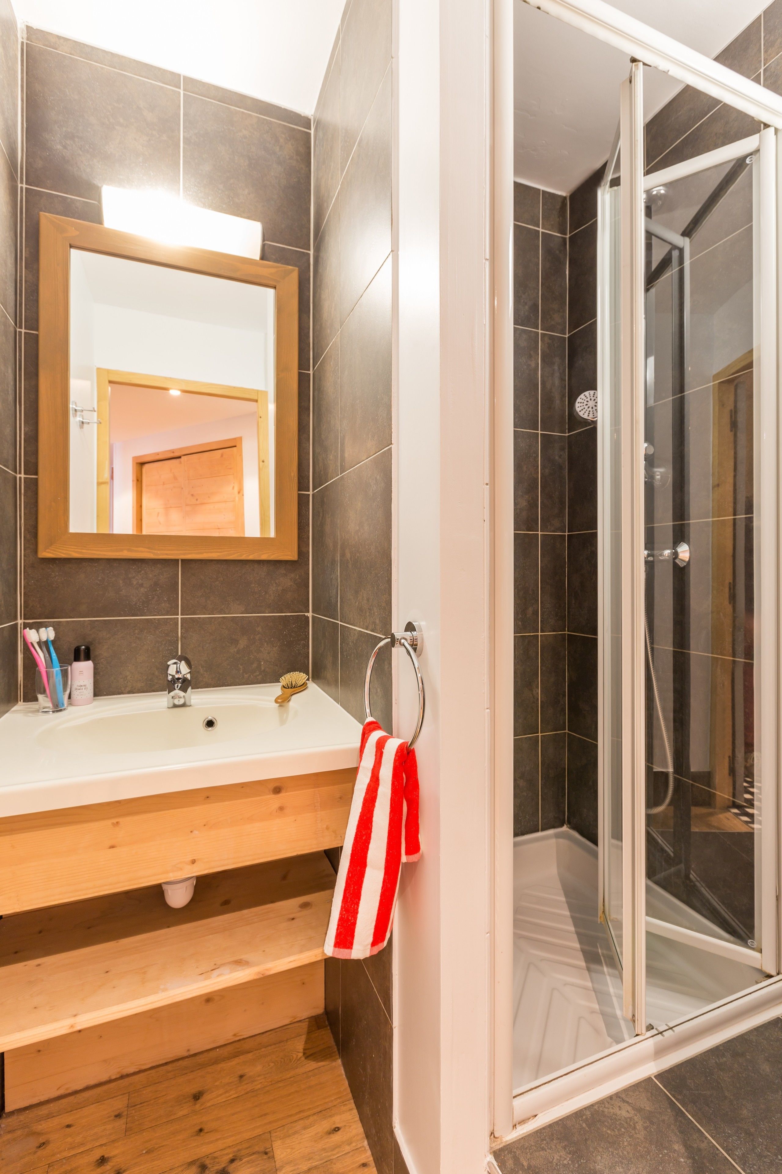 Ein Badezimmer mit einem Holzbadezimmerhocker, einem Spiegel, einer Dusche mit Glastüren und einem rot-weißen gestreiften Handtuch.