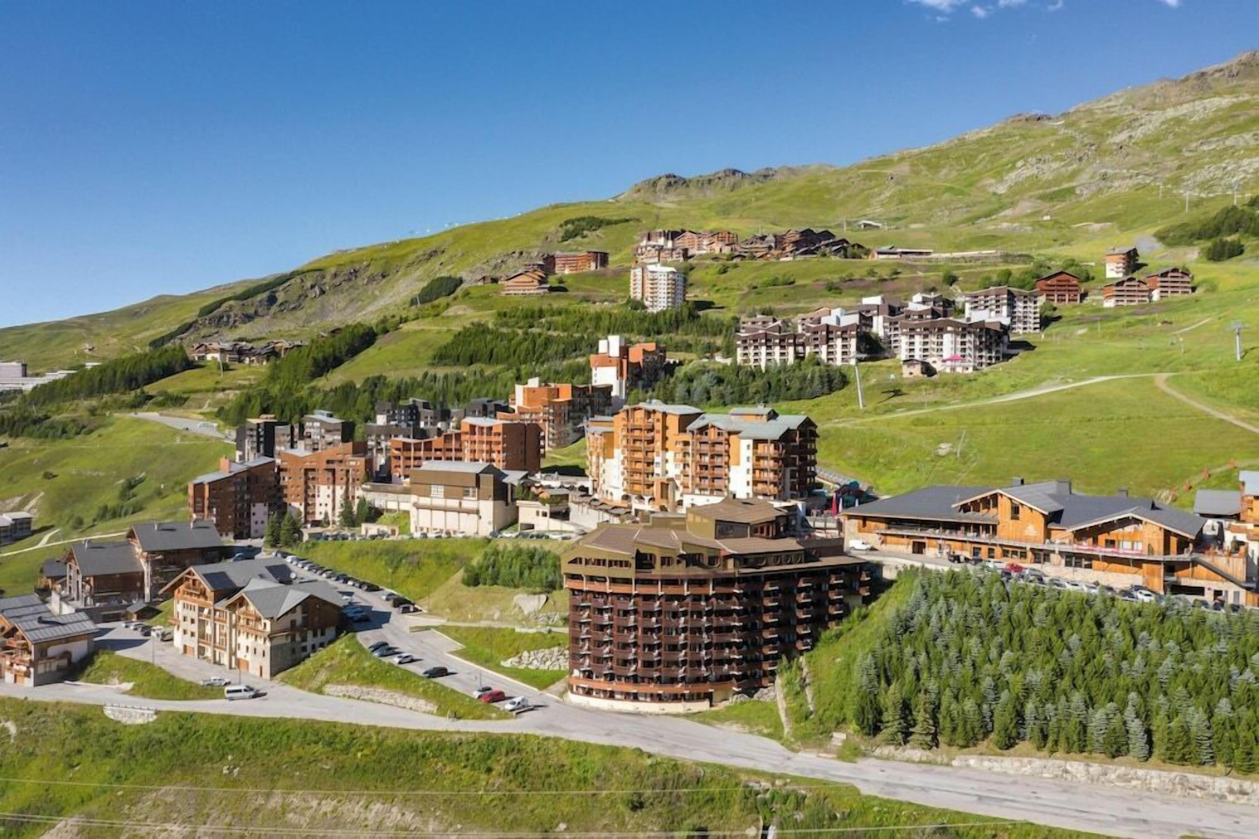 Zona turística montañosa con varios edificios de alojamiento, incluyendo un hotel grande de varios pisos con balcones, rodeada de vegetación y un cielo despejado.
