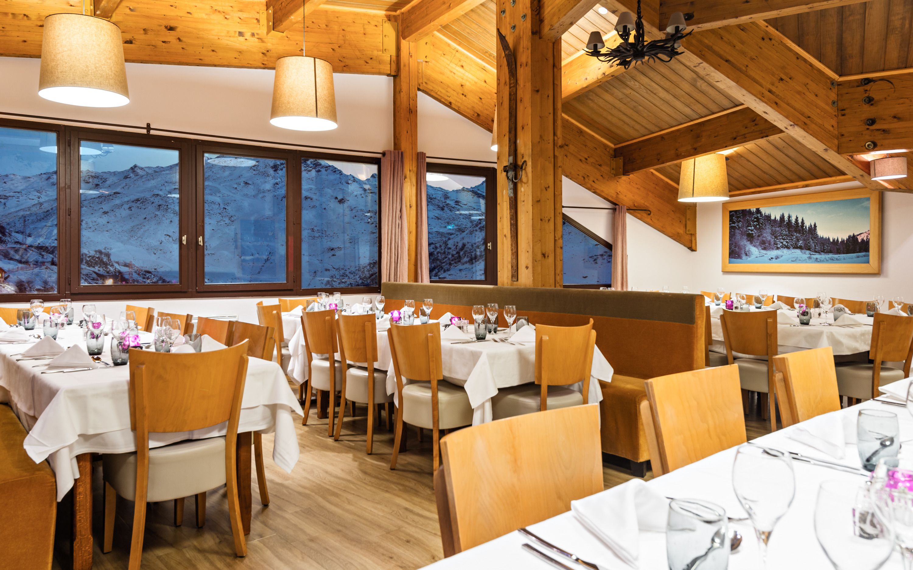 En restaurant med træinteriør, store vinduer med udsigt til bjerge, og borde dækket til spisning.
