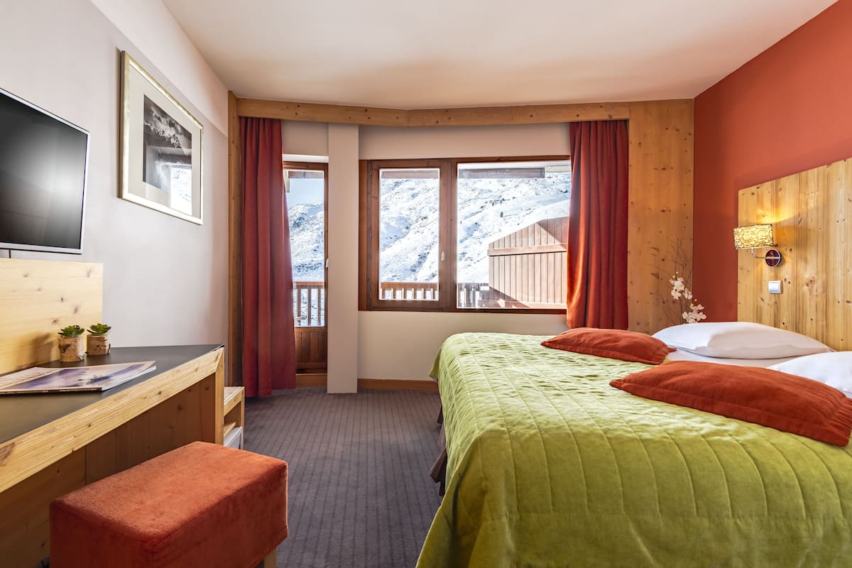 Quarto de hotel com cama, escrivaninha, televisão e janela com vista para uma montanha nevada.