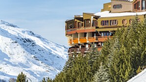 Un edificio de varios pisos con balcones, rodeado de montañas nevadas y árboles de hoja perenne.