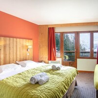 A hotel room with a wooden headboard, a green bedspread, two rolled towels, and a window with a view of trees.