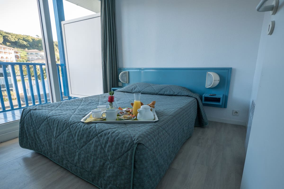 A hotel room with a bed, a tray with breakfast items, a bedside table with a lamp, and a view of the outside.