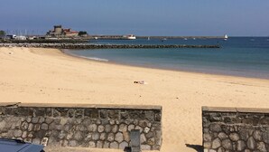 A sandy beach with a stone wall in the foreground, a pier with boats, and a building with a red roof in the background. - Résidence Fort Socoa (Urrugne)