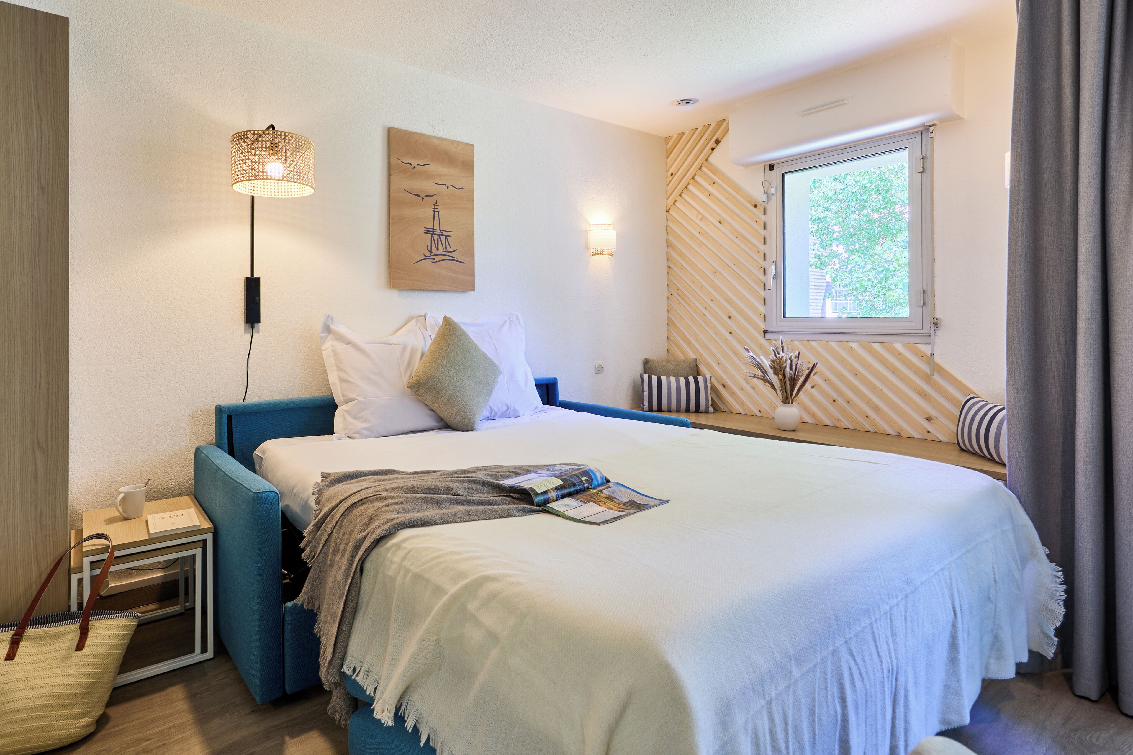 A bedroom with a bed, bedside table, lamp, artwork, window, and curtains.