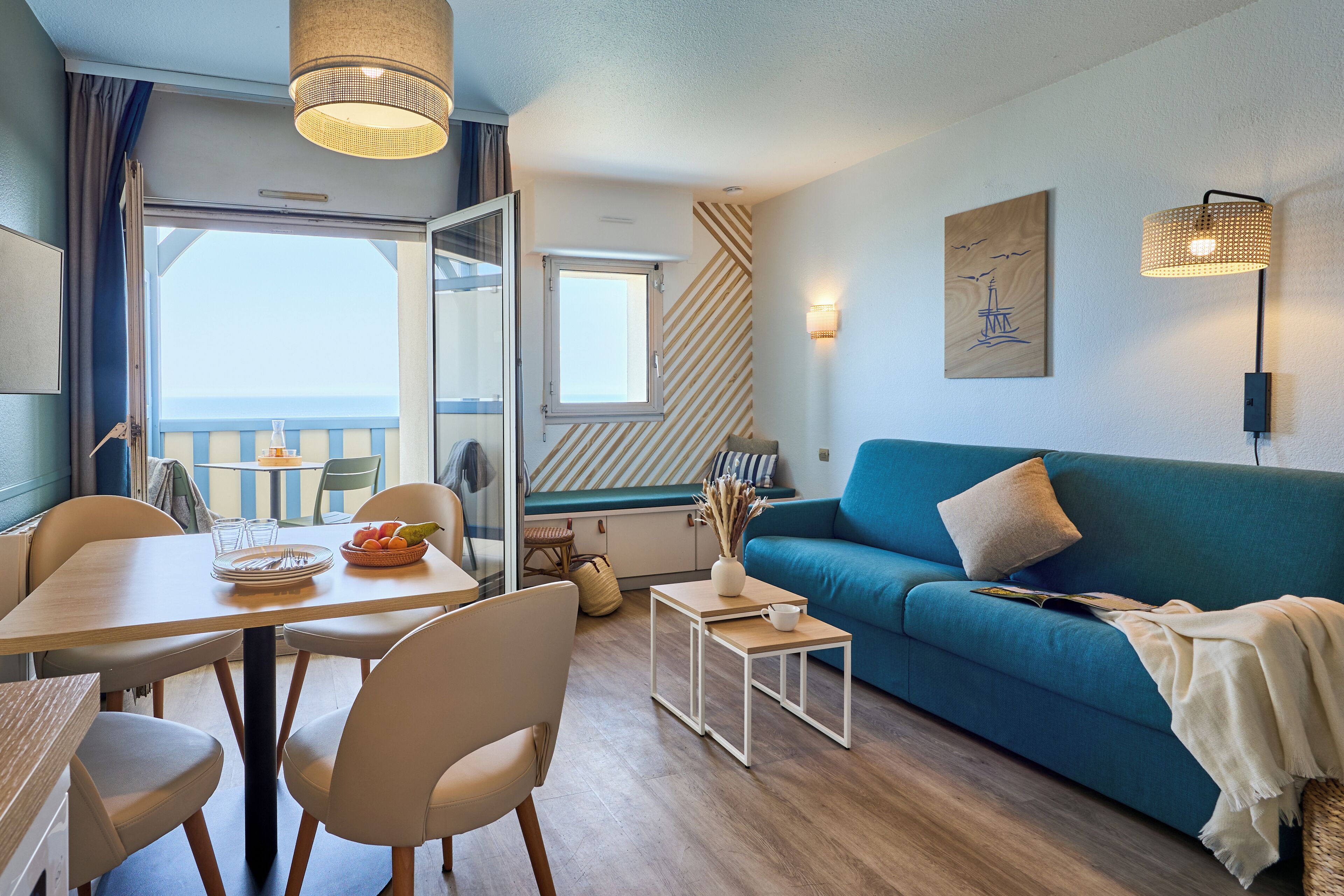 Un salon moderne avec un canapé bleu, une table à manger avec des chaises, une petite table d’appoint et une vue sur la mer par une grande fenêtre.