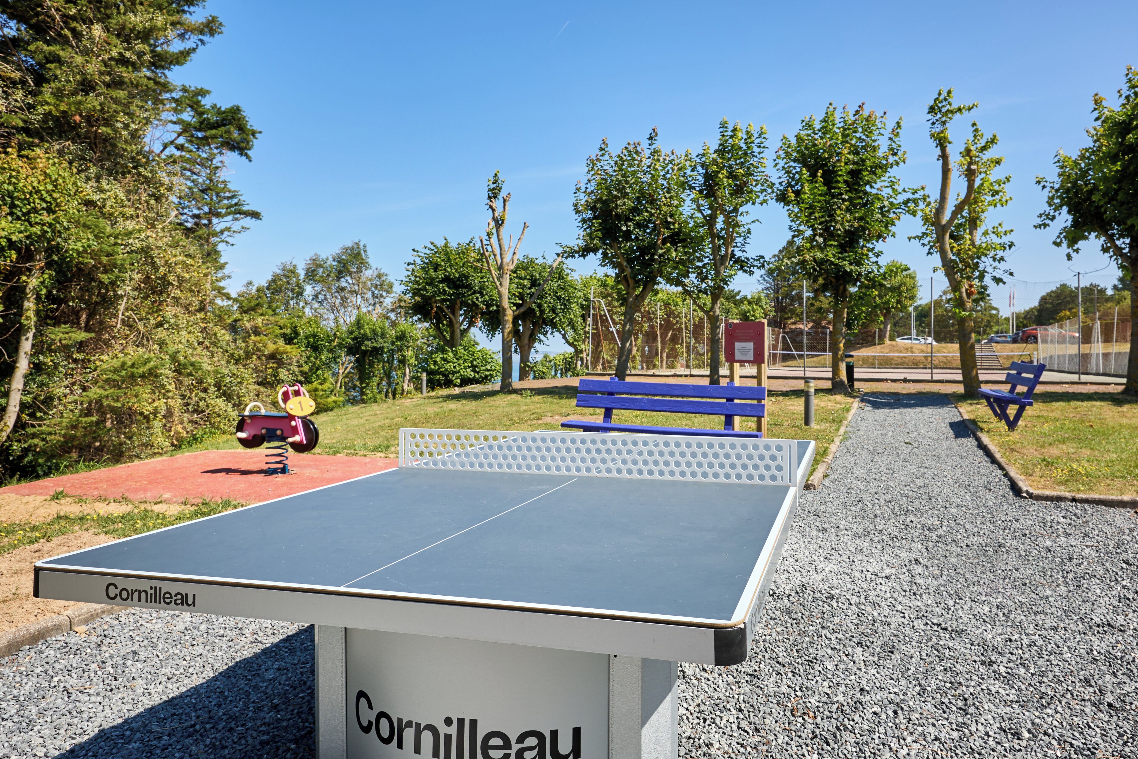 Une table de ping-pong de la marque « Cornilleau » dans un parc, avec des bancs et des arbres.