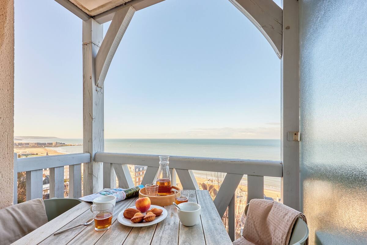 A wooden table with a bottle, glasses, and food on a balcony overlooking the sea.