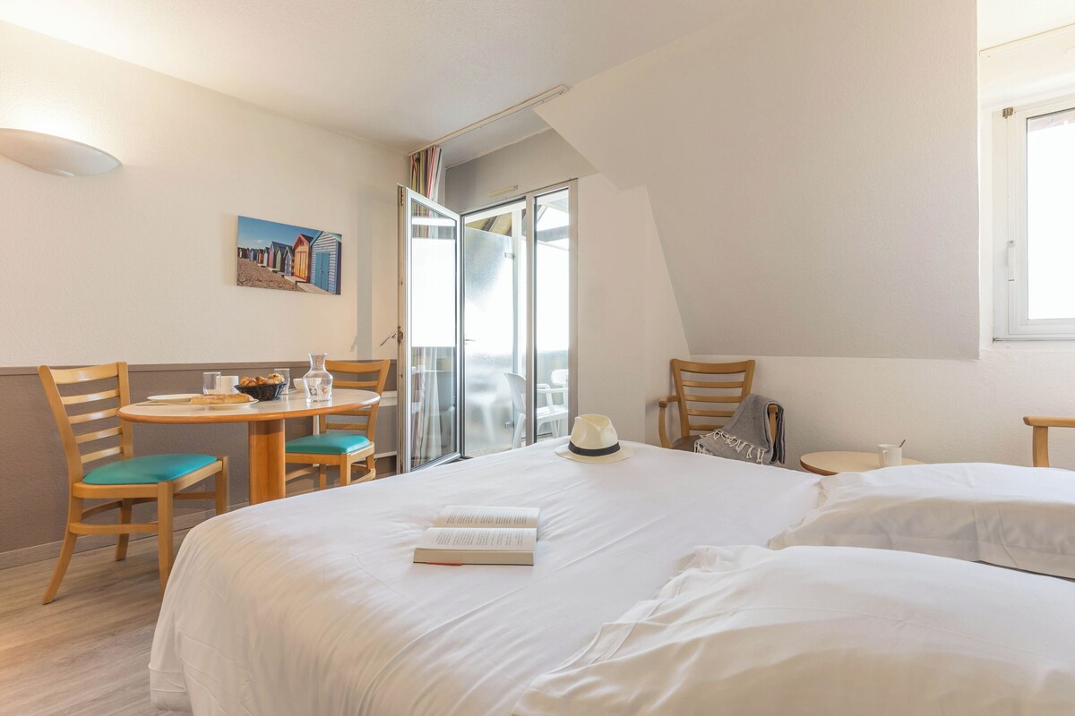 Habitación de hotel con cama, una mesa de comedor con sillas, una mesita con una taza y un cuadro en la pared.