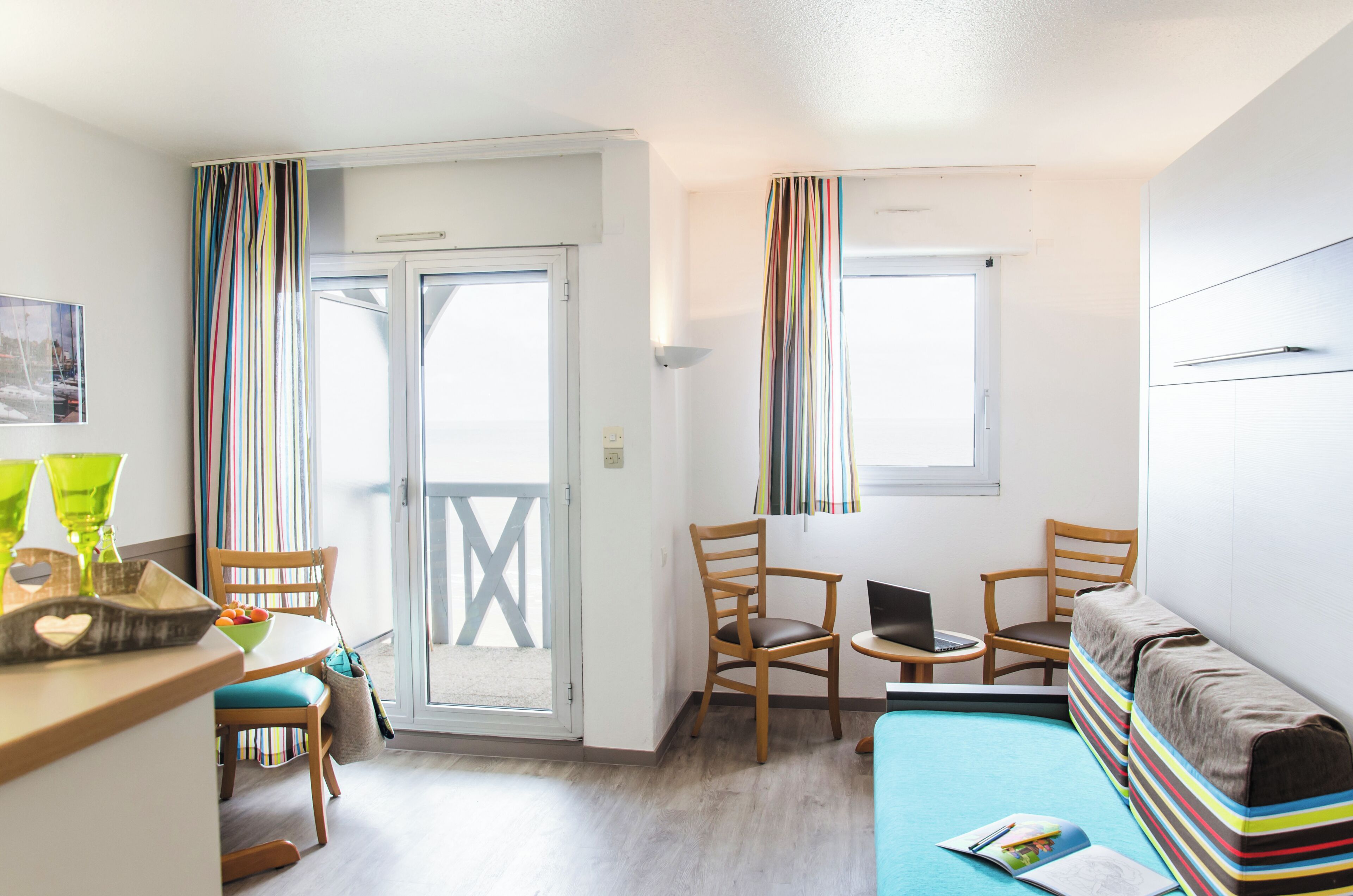 Una habitación con una mesa de comedor, sillas, un ordenador portátil sobre una mesita, un sofá y una ventana con cortinas.