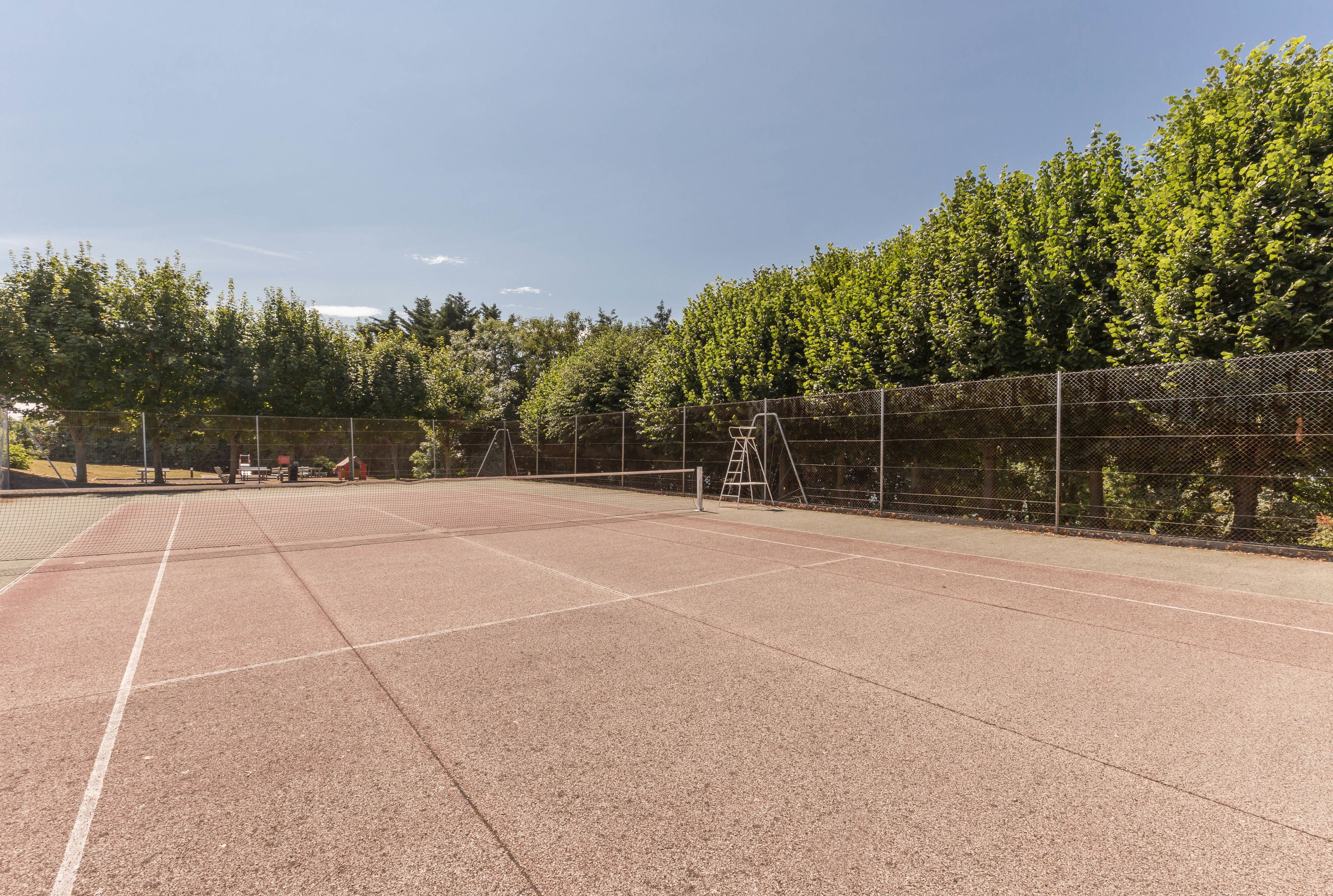Un campo da tennis all'aperto con una rete, circondato da una recinzione e alberi.