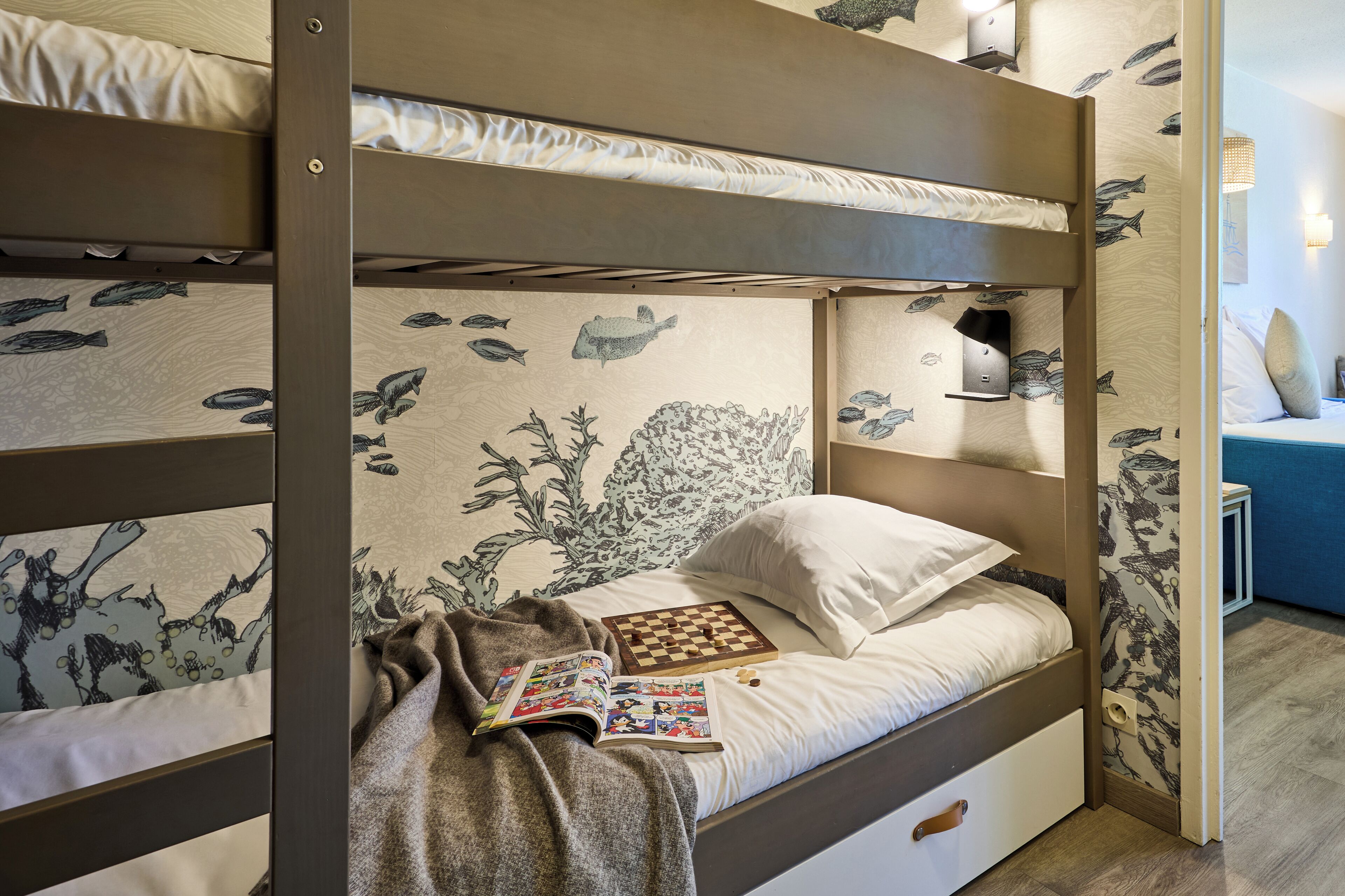 Un lit superposé avec un mur décoré sur le thème des poissons, une petite étagère et un livre sur le lit du bas.