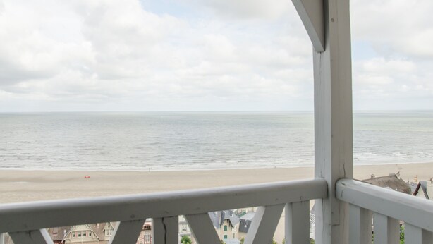Vista de la playa desde un balcón con barandilla, océano y edificios al fondo.