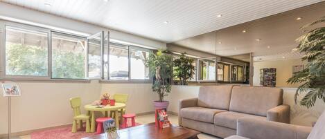 A modern living room with a wooden coffee table, beige sofa, and green chairs. There is a large window with a view of trees outside.