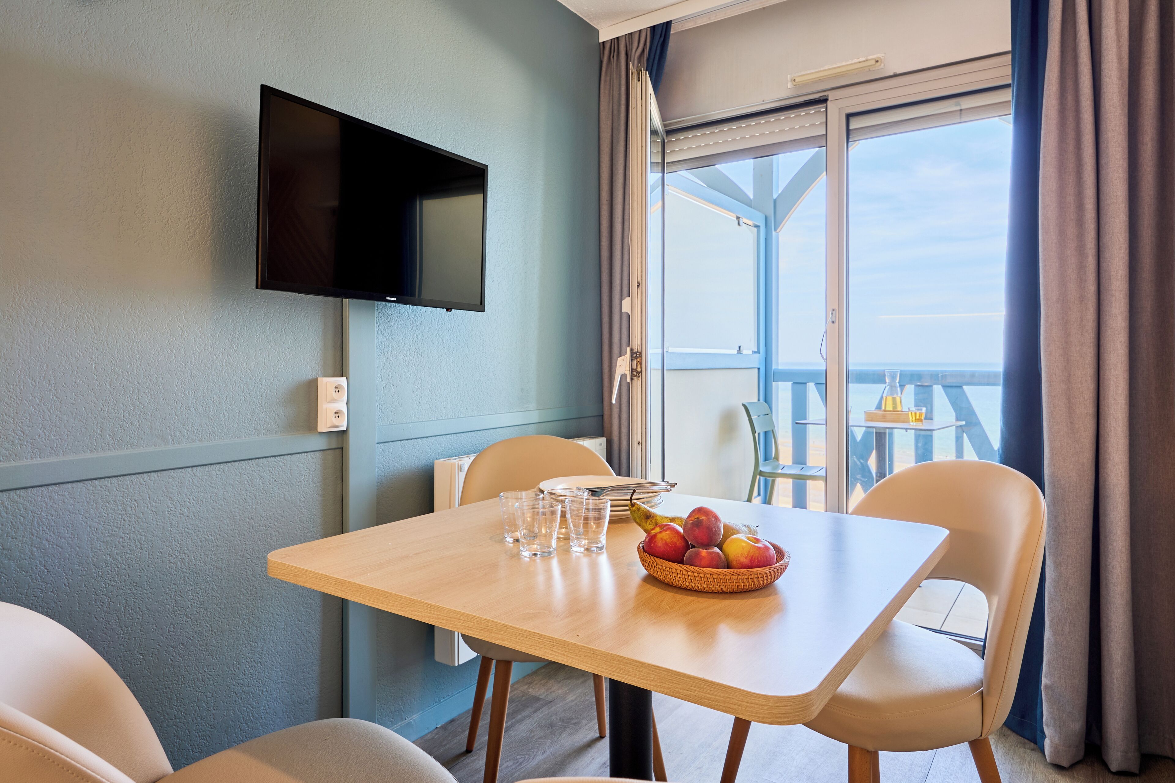 A room with a table, chairs, a television mounted on the wall, and a view of the sea through a balcony.