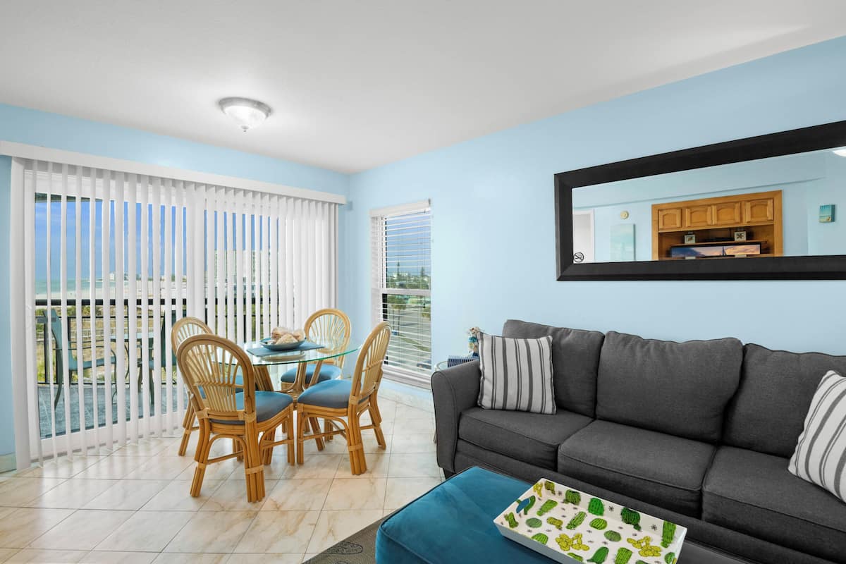 A living room with a sofa, a dining table with chairs, a balcony with white blinds, and a kitchen area visible through a window.