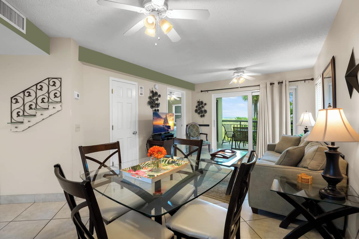 A dining area with a glass table, chairs, a ceiling fan, and a view of the outdoors.