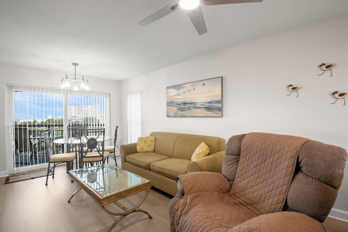 A living room with a glass coffee table, a brown armchair, a beige sofa, a dining area with metal chairs, and a ceiling fan.
